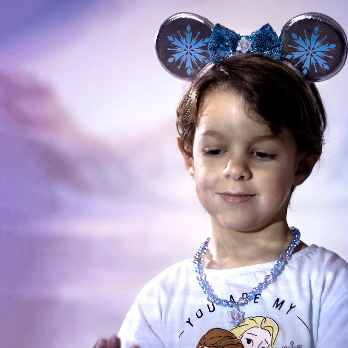 Child wearing a Frozen-themed necklace and headband with snowflake ears against a soft background.